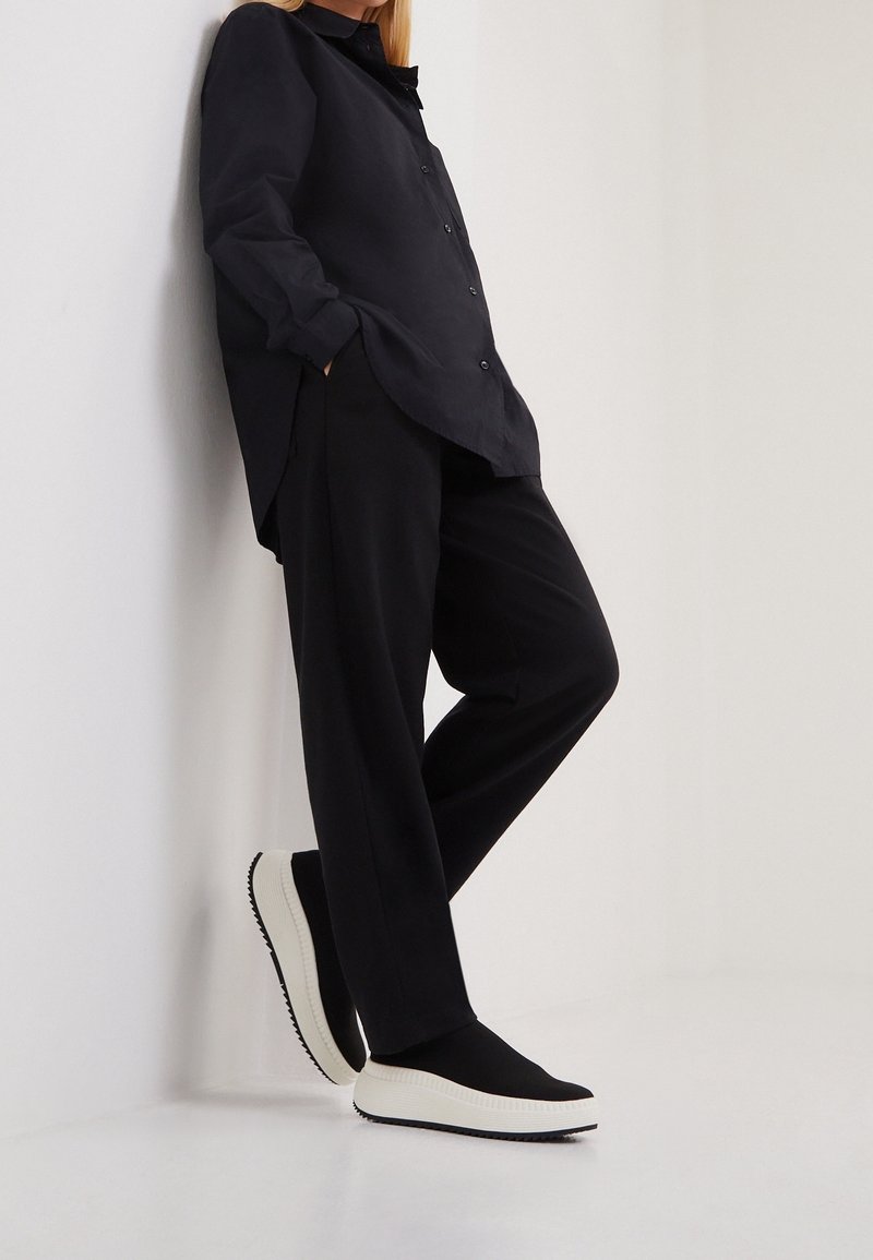 Personne appuyée contre un mur portant un pantalon noir ample, une chemise noire oversize boutonnée et des chaussures noires à enfiler avec des semelles blanches.