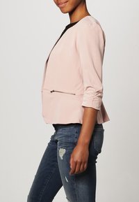 Vaaleanpunainen bleiseri, jossa on sileä pinta, jämäkkä muotoilu, kolmanneksittaiset hihat ja vetoketjullinen sivutasku; yhdistettyinä kuluneisiin farkkuihin.