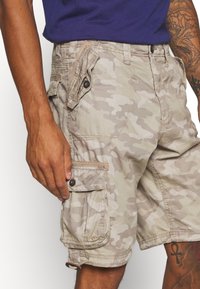 Camouflage cargo shorts in light beige with subtle gray patterns, featuring multiple pockets, a button closure, and durable fabric.