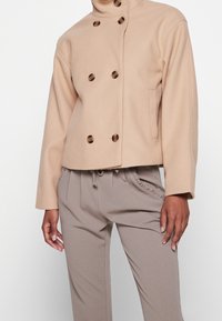 Femme portant une veste en laine beige croisée à boutons écaille de tortue et un pantalon taille haute taupe avec bordure de poche à volants.