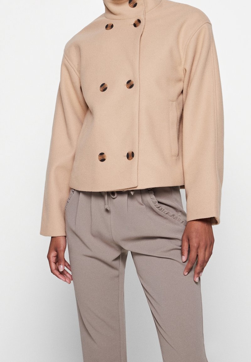 Femme portant une veste en laine beige croisée à boutons écaille de tortue et un pantalon taille haute taupe avec bordure de poche à volants.
