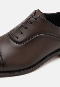 Scarpa da uomo in pelle marrone con finitura liscia, sistema di allacciatura chiusa, punta arrotondata e dettagli di cucitura sottili lungo la tomaia e la suola.