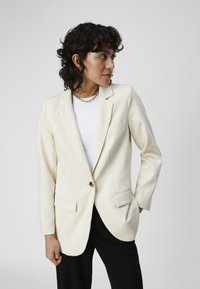 Blazer ajusté blanc cassé en tissu doux, fermeture à un bouton, deux poches avant, revers crantés, associé à un haut blanc et un pantalon noir.