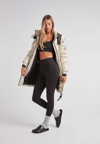 Beige pufferjack met zwarte accenten, gecombineerd met zwarte leggings, grijze sokken en zwarte slippers; heeft een ritsluiting en binnenzakken.