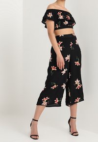 Svart off-shoulder crop top med blommig broderi, matchad med avkortade blommiga byxor med en lös design, förhöjd av svarta klackar.