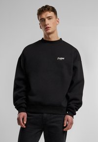 Czarny sweatshirt z okrągłym dekoltem i luźnym krojem. Na przodzie haftowane słowo "Zabronione" w białym kolorze. Miękka, teksturowana tkanina.