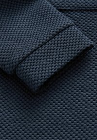 Geweven donkerblauwe stof met een gestructureerd patroon en een opgerolde manchet. Het materiaal lijkt stevig en tastbaar, met een rasterachtig ontwerp.
