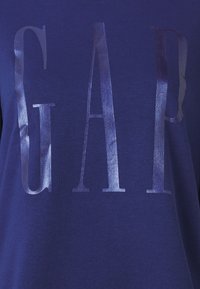 Camiseta de algodón azul marino con letras "GAP" plateadas brillantes. Textura suave, mangas cortas, diseño de cuello redondo y un corte relajado.
