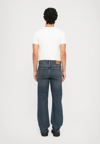 Jean en denim bleu avec une coupe droite, poches arrière et une étiquette en cuir beige à la taille. Associé à un T-shirt blanc uni.