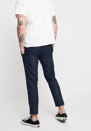 Marineblå bukser med nålestriber i letvægtsstof, tætsiddende pasform, kombineret med sorte sneakers og en hvid kortærmet skjorte.