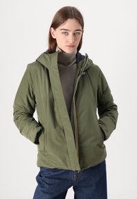 Giacca a vento verde oliva con cappuccio e chiusura zip, caratterizzata da un esterno testurizzato e tasche laterali, indossata sopra un top aderente marrone e jeans blu.