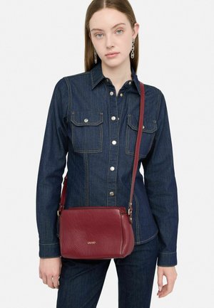 Borsa a tracolla rossa in pelle con finitura testurizzata, tracolla regolabile e un accento del logo dorato. Indossata con una camicia di jeans e jeans blu.