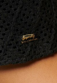 Textura de punto negro con un patrón de punto abierto. Etiqueta dorada "Superdry Spirit of Adventure" se exhibe de manera prominente.
