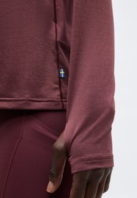 Μπλούζα μακρυμάνικη σε βαθύ μπορντό χρώμα με υφή και τρύπα για τον αντίχειρα στο μανίκι, φορεμένη με αντίστοιχο κολάν, με μικρή ταμπέλα με τη σημαία της Σουηδίας κοντά στο τελείωμα.