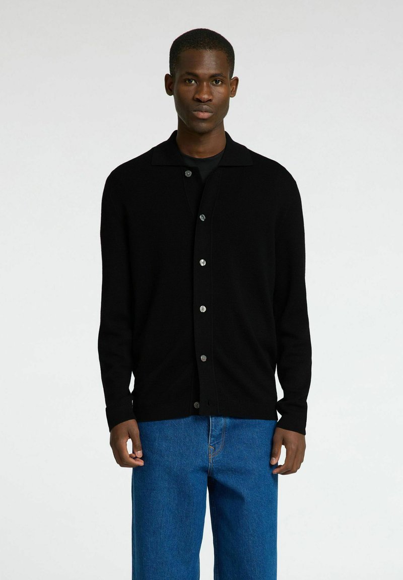 Svart cardigan med spiss krage, lange ermer og knapper foran. Brukt over en svart skjorte og kombinert med blå vide jeans.