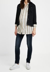 Svart denimjacka över en vit och beige randig skjorta med knappar, tillsammans med mörka skinny jeans och vita sneakers.