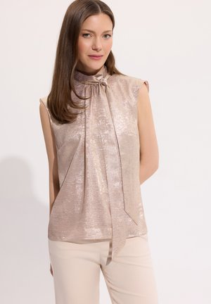 Kobieta o prostych brązowych włosach, ubrana w błyszczącą beżową bluzkę bez rękawów z wysokim kołnierzem i długim wiązaniem z przodu, w połączeniu z jasnobeżowymi spodniami.