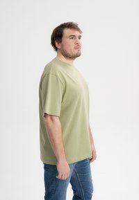 Olivgrünes T-Shirt mit kurzen Ärmeln und Rundhalsausschnitt, aus weichem Stoff; kombiniert mit blauen Jeans im lässigen Schnitt.