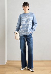 Lys blå strikkegenser med hvit grafisk tekst, kombinert med mørk blå flagrende jeans og sølv høye sko. Har en teksturert stoff.