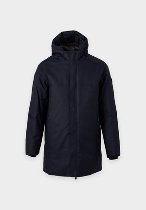 Un homme porte un manteau bleu marine à capuche par-dessus un pull crème, un pantalon beige et des bottes marron, sur un fond clair et uni.