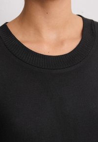 Primo piano del collo e della parte superiore del petto di una persona che indossa un maglione nero a collo rotondo con dettagli a coste sul colletto.