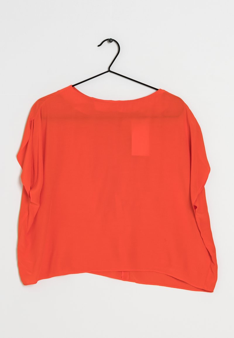 Blouse rouge, transparente, à manches courtes avec un col large et une coupe ample. L'ourlet est arrondi à l'avant et plus long à l'arrière. Accrochée à un crochet noir.