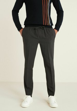 Homme portant un pull noir à rayures verticales, un pantalon gris foncé avec cordon de serrage et des baskets blanches, debout les mains dans les poches.