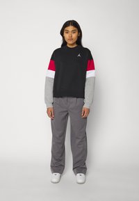 Sudadera negra con mangas en rojo, blanco y gris, que presenta un pequeño logo. Combinada con pantalones grises a rayas y zapatillas blancas.