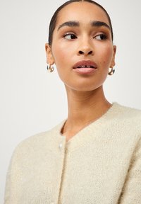 Cardigan en maille beige avec une texture duveteuse, col rond et fermeture à boutons en perles. Le modèle porte des boucles d'oreilles créoles en argent.