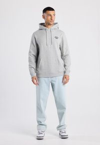 Felpa grigia con cappuccio, tasca frontale e logo stampato. Abbinata a jeans azzurri chiari e sneakers bianche.