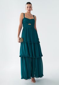 Abito lungo turchese con corpetto aderente, spalline sottili e dettaglio a taglio. Presenta balze sovrapposte all'orlo. La modella tiene in mano una pochette dorata.