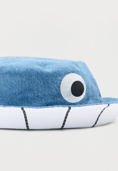 Μπλε τζιν καπέλο με στρογγυλεμένο σχήμα, που διαθέτει ένα μεγάλο λευκό μάτι με μαύρη κόρη. Το λευκό γείσο έχει μαύρες γραμμές και υφή επιφάνειας.