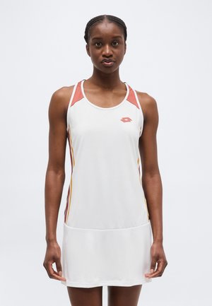 Žena v bílých sportovních šatech bez rukávů s rezavými a hořčicovými akcenty, stojící před jednoduchým světlým pozadím.