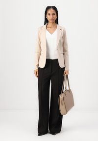 Blazer beige sopra un top bianco a scollo a V, abbinato a pantaloni neri a gamba larga. Tiene una borsa color cammello. Tessuto dalla superficie liscia, vestibilità sartoriale.