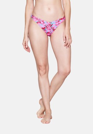 Vrouw draagt roze bikinibroekje met rood- en blauw bloemmotief, staat blootsvoets met gekruiste benen tegen een effen achtergrond.