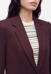 Femme portant un blazer bordeaux foncé sur une chemise blanche avec de fines rayures horizontales noires.