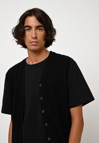 Cardigan noir à manches courtes, en matériau doux, avec des boutons sur le devant et une coupe décontractée, porté sur une chemise noire.