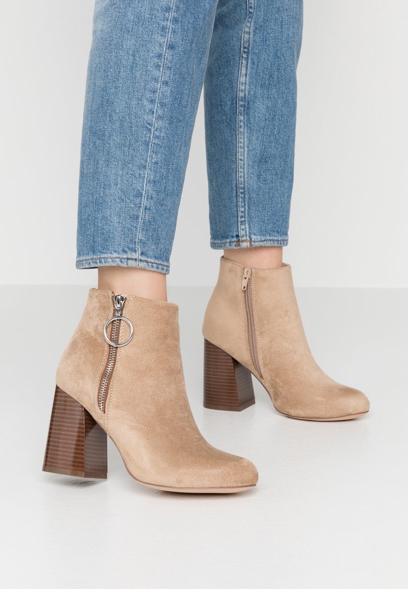 Bottines en daim beige avec un bout rond, fermeture éclair avec un tire-zip circulaire, et un talon épais marron texturé.