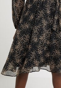 Svart klänning med korta puffärmar, som har ett strukturerat tyg och ett spritt mönster av guldprickar. Nederkanten når till midjan på vaden.