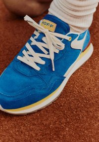 Niebieskie zamszowe sneakersy z białymi sznurówkami, ż�ółtymi akcentami i teksturowanym wykończeniem. Posiadają białe logo i charakterystyczny wzór podeszwy.