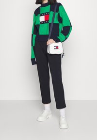 Grün und marineblau karierter Pullover, schwarze Hose, weiße Schuhe und eine kleine weiße Tasche mit Logo. Strukturierter Strickstoff und lockere Passform.