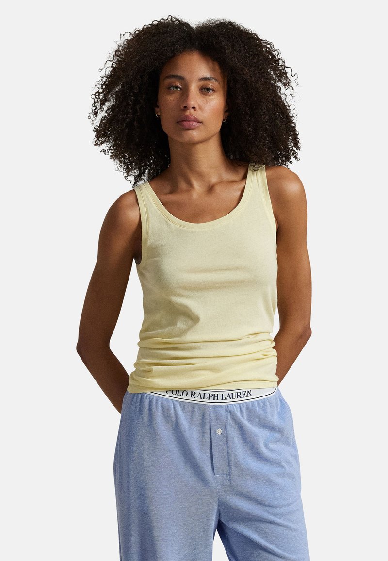 Femme aux cheveux bouclés portant un haut jaune pâle sans manches et un pantalon bleu clair avec une ceinture Polo Ralph Lauren, debout devant un fond blanc.