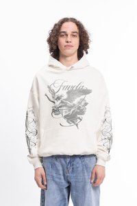 Krémová mikina s kapucňou a grafickým dizajnom, ktorý zobrazuje anjela a vírive vzory. Má predné vrecko a rebrované manžety. Nosená s modrými džínsami.