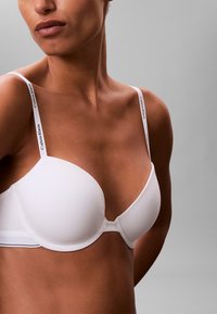 Kobieta nosząca biały biustonosz Calvin Klein z markowymi ramiączkami na gołej skórze na jednolitym szarym tle.