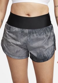 Personne portant une brassière de sport blanche et un short de sport gris taille haute avec une ceinture noire, debout, les bras détendus le long du corps.