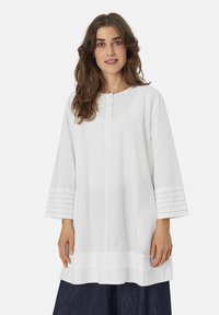 Witte katoenen tuniek met een ronde halslijn en knoopsluiting. Heeft driekwartmouwen met oogjes en een rechte snit.