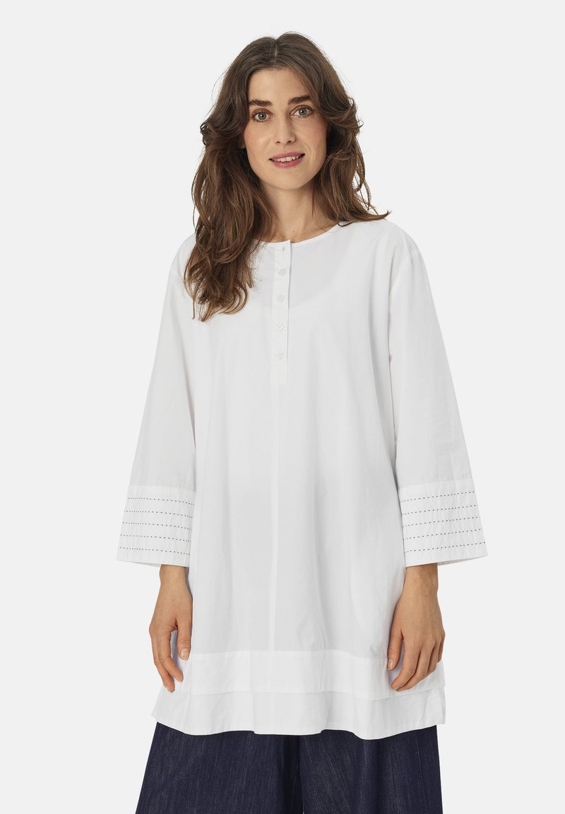 Witte katoenen tuniek met een ronde halslijn en knoopsluiting. Heeft driekwartmouwen met oogjes en een rechte snit.