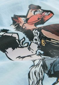 Tricou cu un imprimeu colorat de benzi desenate, având personaje în albastru și roșu, pe un material de culoare albastru deschis. Textura pare netedă și sintetică.