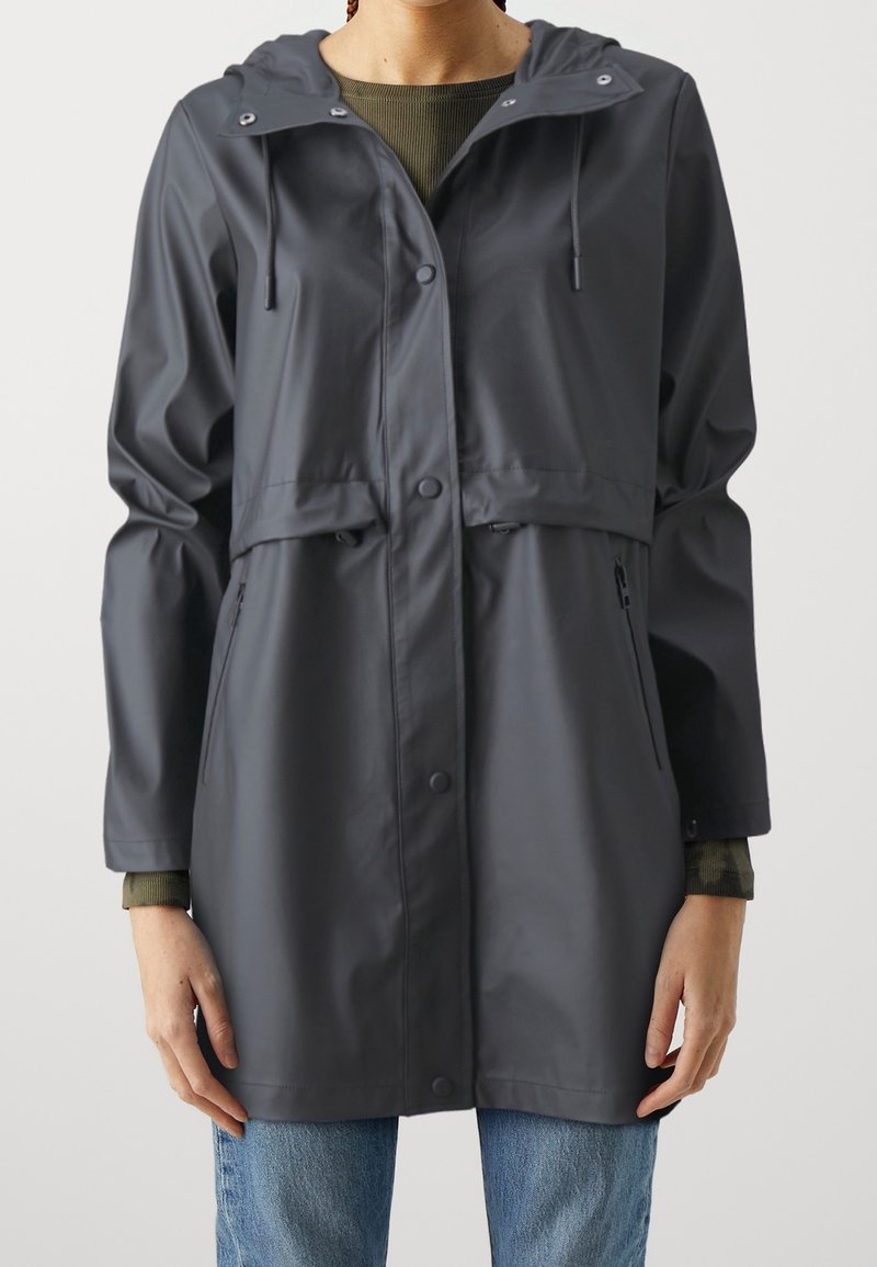 Personne portant un imperméable à capuche gris foncé avec des boutons-pression et des poches zippées sur un haut vert et un jean bleu.