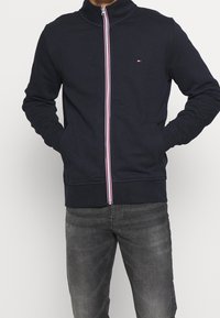 Granatowa bluza rozpinana na zamek z wysokim kołnierzem oraz kontrastowym zamkiem w czerwono-białe paski. Posiada dwie boczne kieszenie i małe logo na piersi.